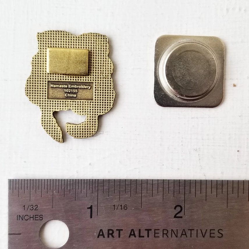The Otterly Adorable Enamel Needle Minder by Jessica Long Embroidery features a magnetic pin with a rectangular clasp and square metal backing, shown next to an inch ruler on a white surface.