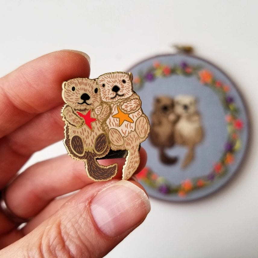 A hand holds the Otterly Adorable Enamel Needle Minder by Jessica Long Embroidery, featuring two hugging otters with stars. In the blurred background is matching otter embroidery surrounded by a floral wreath.