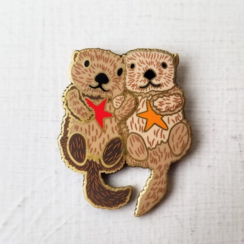 The Otterly Adorable Enamel Needle Minder by Jessica Long Embroidery features two illustrated sea otters holding hands and starfish—one red, one orange—against a plain white background. Perfect for keeping track of your needles!.