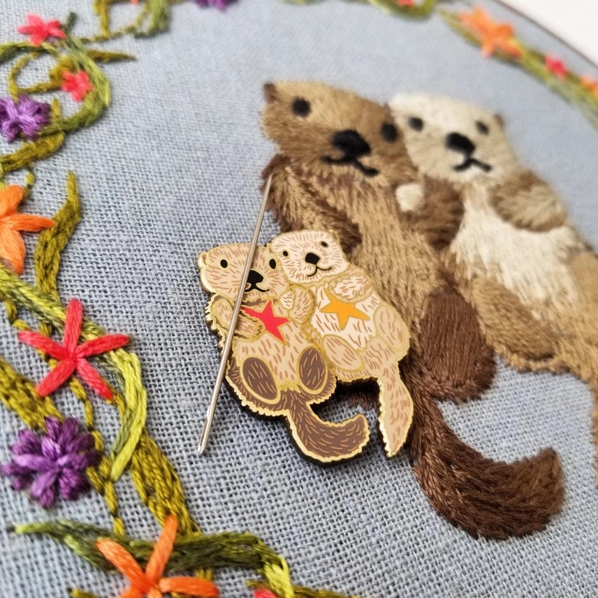 A close-up of embroidery art featuring two otters with floral accents, topped with the Otterly Adorable Enamel Needle Minder by Jessica Long Embroidery, shaped like hugging sea otters and holding a sewing needle.