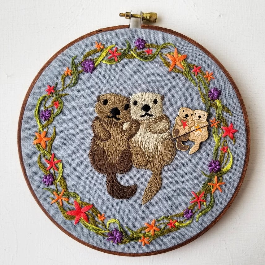 The Otterly Adorable Enamel Needle Minder by Jessica Long Embroidery features two hand-embroidered otters holding hands in a floral wreath, with a mini hard enamel otter as a magnetic needle minder for added convenience.