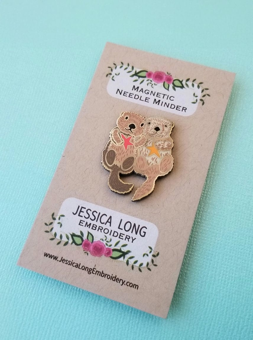 The Otterly Adorable Enamel Needle Minder by Jessica Long Embroidery features two cuddling otters holding a heart, and arrives on a floral card.