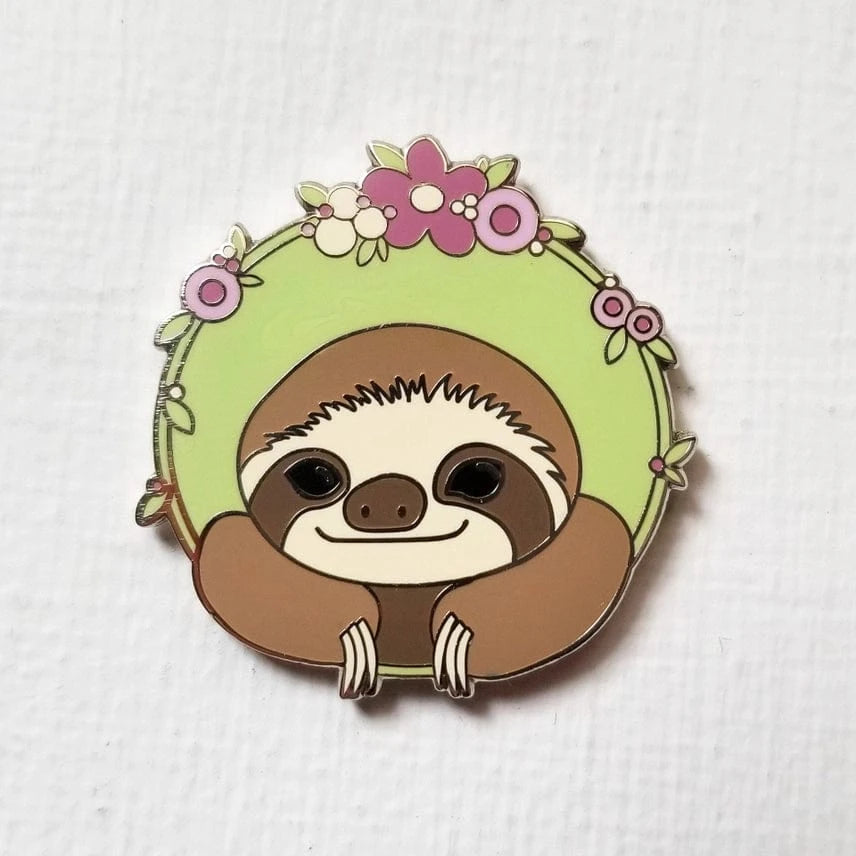 The Sloth Enamel Needle Minder by Jessica Long Embroidery features a smiling sloth resting inside a green circle with pink and purple flowers—crafted in hard enamel and perfect for keeping your needles secure on any project.