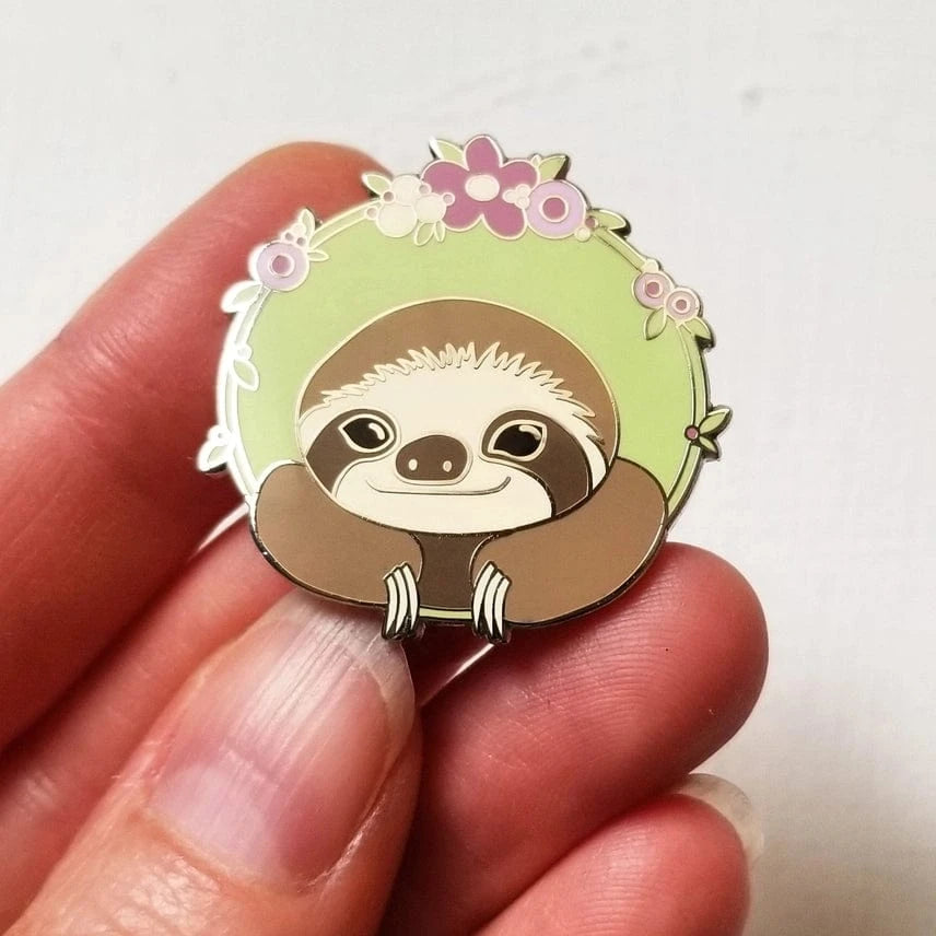 A hand holds the Jessica Long Embroidery Sloth Enamel Needle Minder—a hard enamel pin with a smiling cartoon sloth, green border, and pink and purple flowers—perfect for keeping needles secure during crafts.