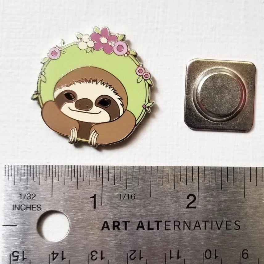 The Sloth Enamel Needle Minder by Jessica Long Embroidery features a cute sloth with a floral wreath, measures about 1.25 inches wide, and sits on a white textured surface next to its backing—a perfect tool for needlework.