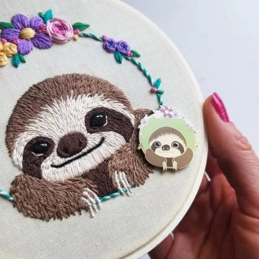 A hand with pink nails holds a floral embroidered sloth hoop by Jessica Long Embroidery, with the Sloth Enamel Needle Minder—a matching magnetic pin—displayed beside the sloth’s face.