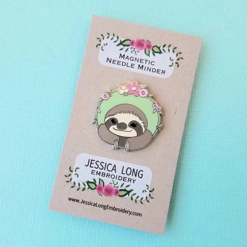 The Sloth Enamel Needle Minder by Jessica Long Embroidery is a cute magnetic hard enamel pin with a mint green floral background, displayed on a branded card atop a light blue surface.