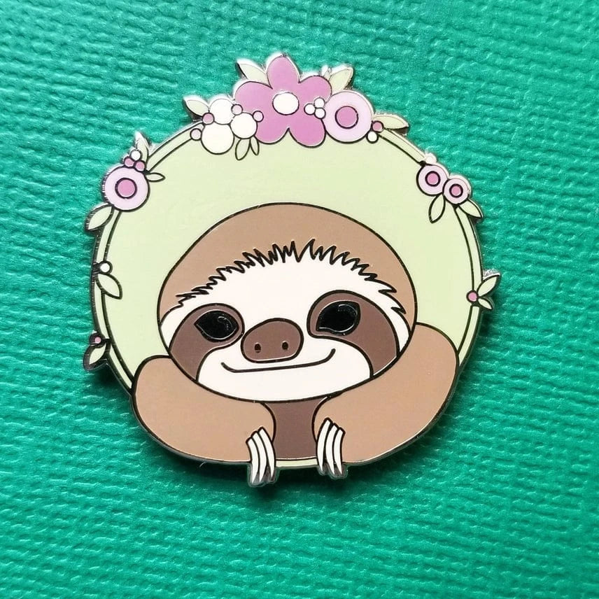 The Sloth Enamel Needle Minder by Jessica Long Embroidery features a smiling sloth with floral accents on pale green—perfect as a magnetic pin for your crafting or embroidery projects.