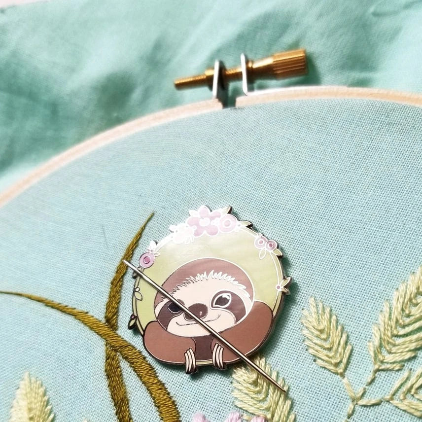 The Sloth Enamel Needle Minder by Jessica Long Embroidery rests on light blue fabric in an embroidery hoop, surrounded by embroidered green leaves and yellow stems.