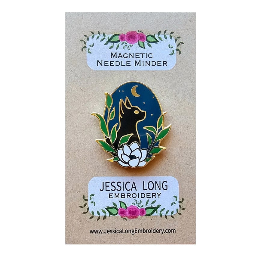 The Stargazer Black Cat Enamel Needle Minder by Jessica Long Embroidery features a hard enamel magnetic pin of a black cat with gold outlines, sitting among green leaves and a white flower beneath a crescent moon.