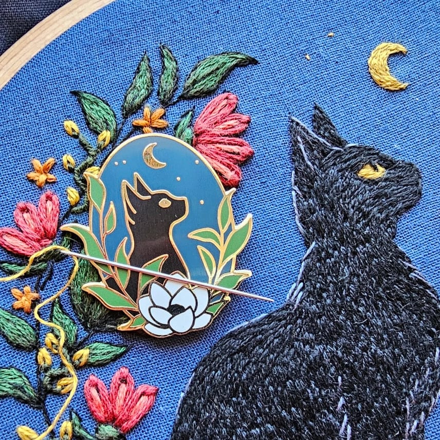 The Stargazer Black Cat Enamel Needle Minder by Jessica Long Embroidery features a black cat with yellow eyes gazing at a crescent moon, echoing the blue floral embroidery design it rests on.