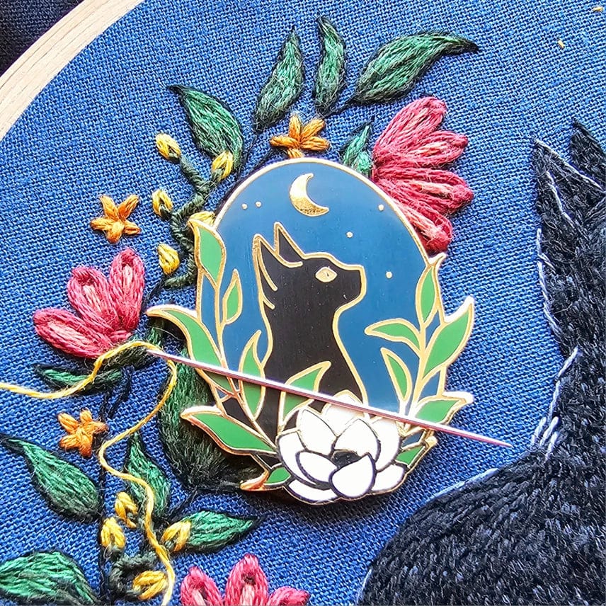 The Stargazer Black Cat Enamel Needle Minder by Jessica Long Embroidery, featuring a moon and leaves, rests on blue fabric with colorful flower embroidery. A needle with red thread lies across this magnetic pin holder.