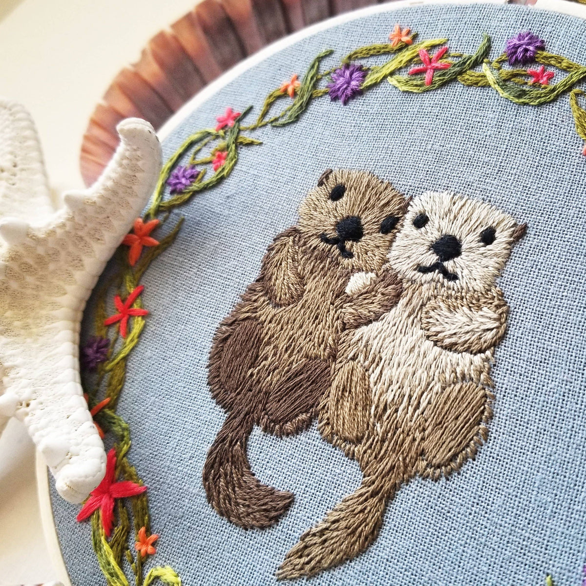 The *Late May Pre-Order* 6 Otterly Adorable Embroidery Kit by Jessica Long Embroidery features two otters holding hands among colorful flowers and green vines—perfect as wall art or a unique handmade gift.