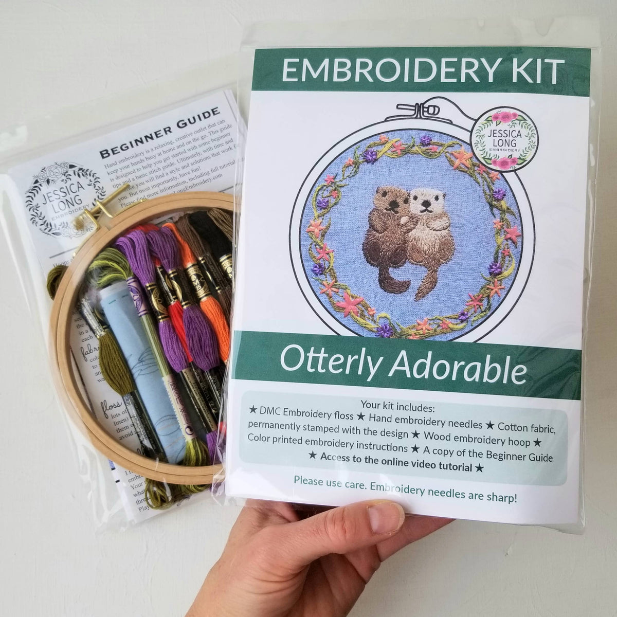 A hand holds the Jessica Long Embroidery *Late May Pre-Order* 6 Otterly Adorable Embroidery Kit, featuring an otter design, vibrant floss, hoop, needles, and a beginner guide—ideal for handmade wall art or gifting, all in clear packaging.