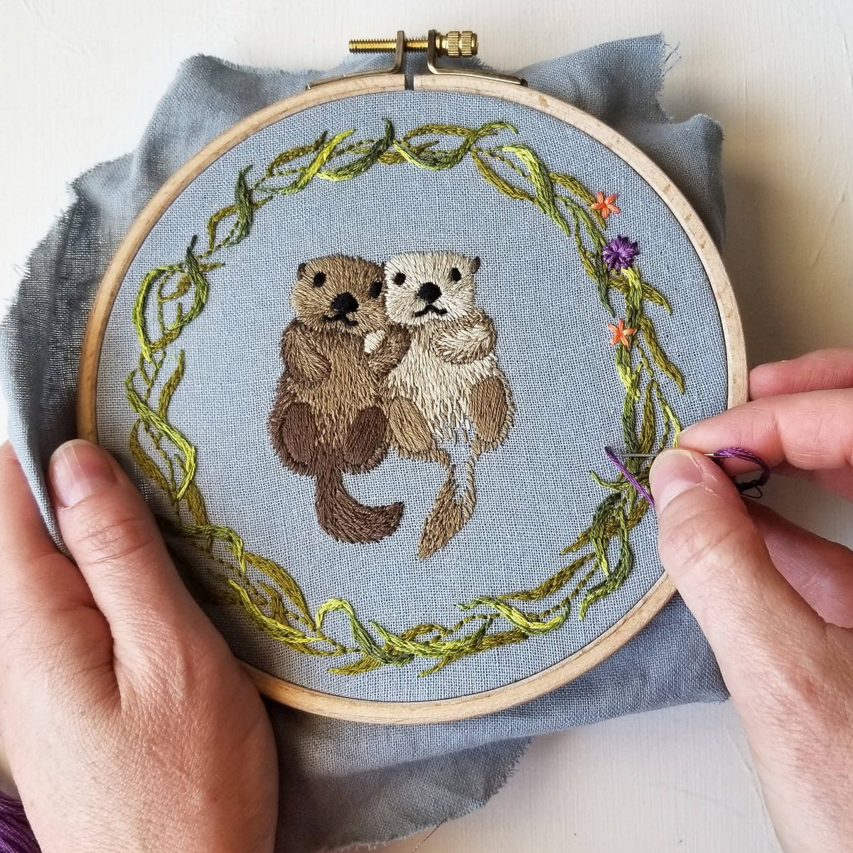 A person holds the *Late May Pre-Order* 6 Otterly Adorable Embroidery Kit by Jessica Long Embroidery, featuring two otters holding hands in a leafy floral wreath on blue fabric—perfect as wall art or a handmade gift.