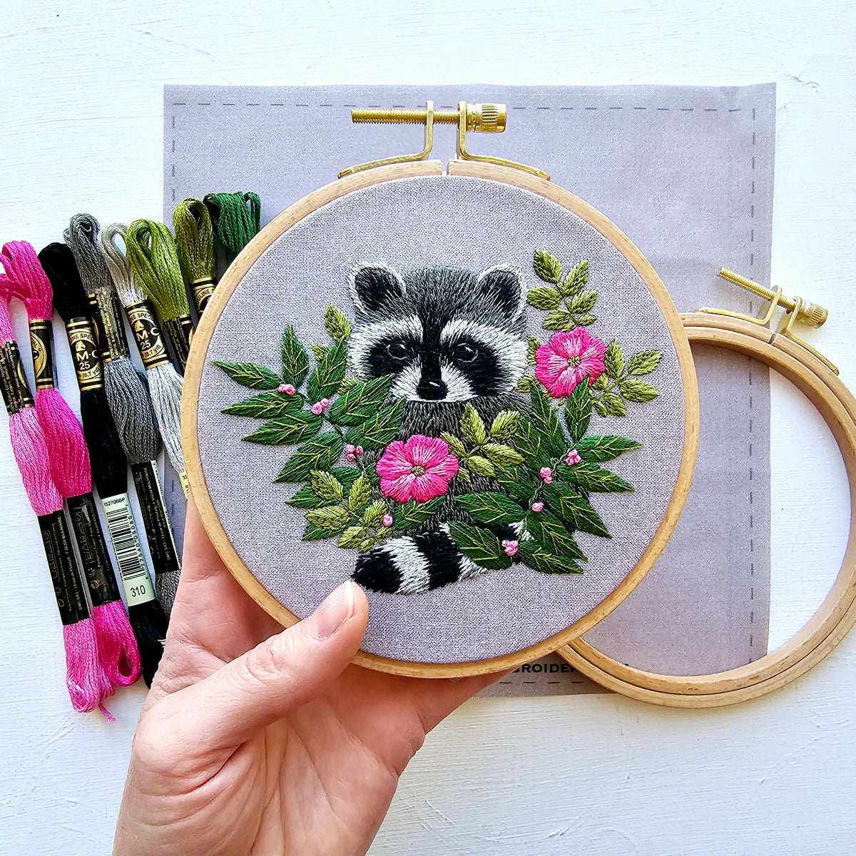 A hand holds Jessica Long Embroiderys 5 Raccoon Embroidery Kit, featuring a raccoon amid pink flowers and green leaves, with pink, green, and black floss ideal for thread painting this charming wildlife craft.