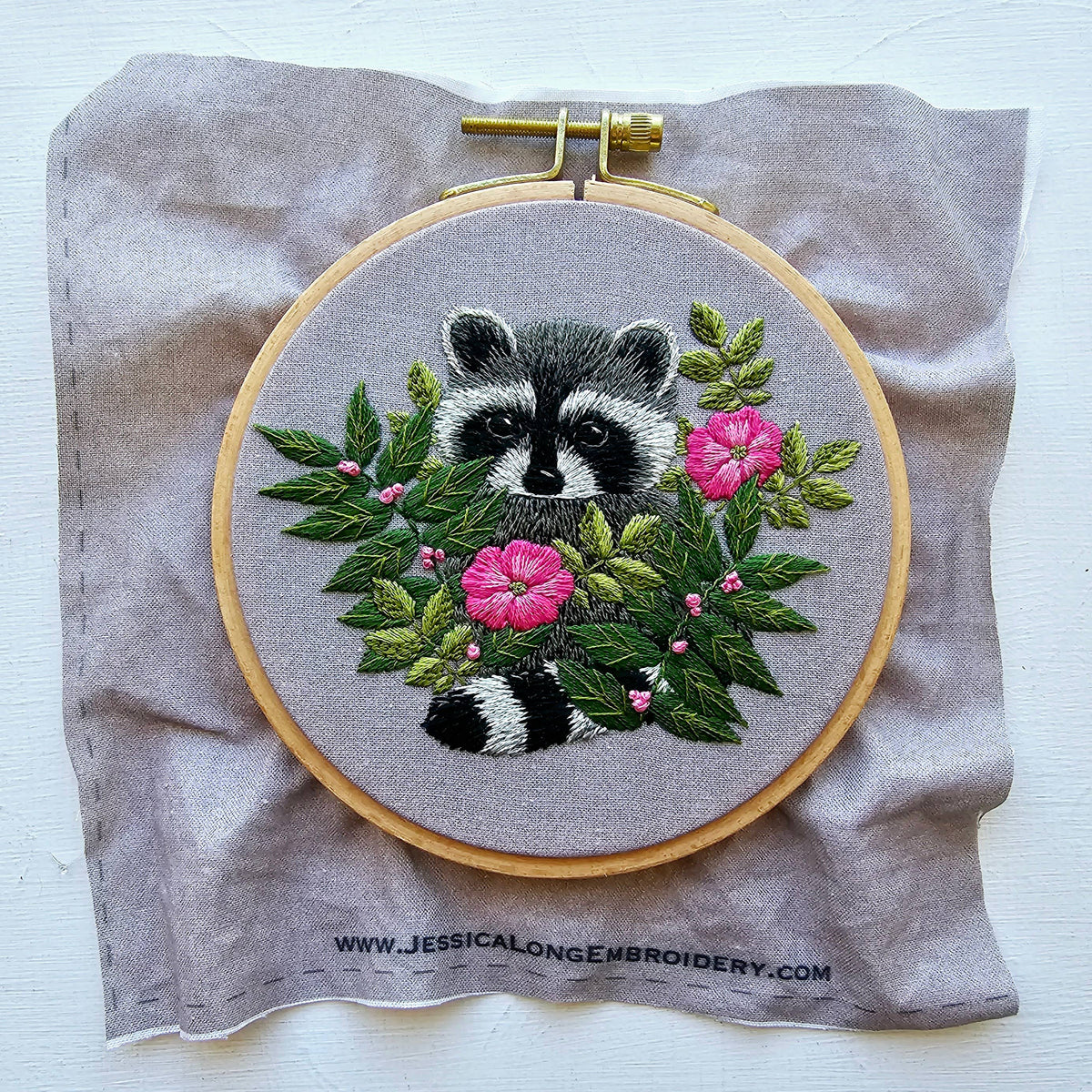 The Jessica Long Embroidery 5 Raccoon Embroidery Kit features a raccoon peeking through leaves and pink flowers, expertly stitched on gray fabric marked with www.JESSICALONGEMBROIDERY.COM at the bottom.