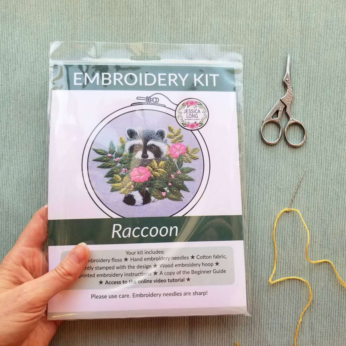 A hand holds the Jessica Long Embroidery 5 Raccoon Embroidery Kit, which features a raccoon with pink flowers on the package. Silver scissors and yellow thread rest nearby on a light green textured surface.