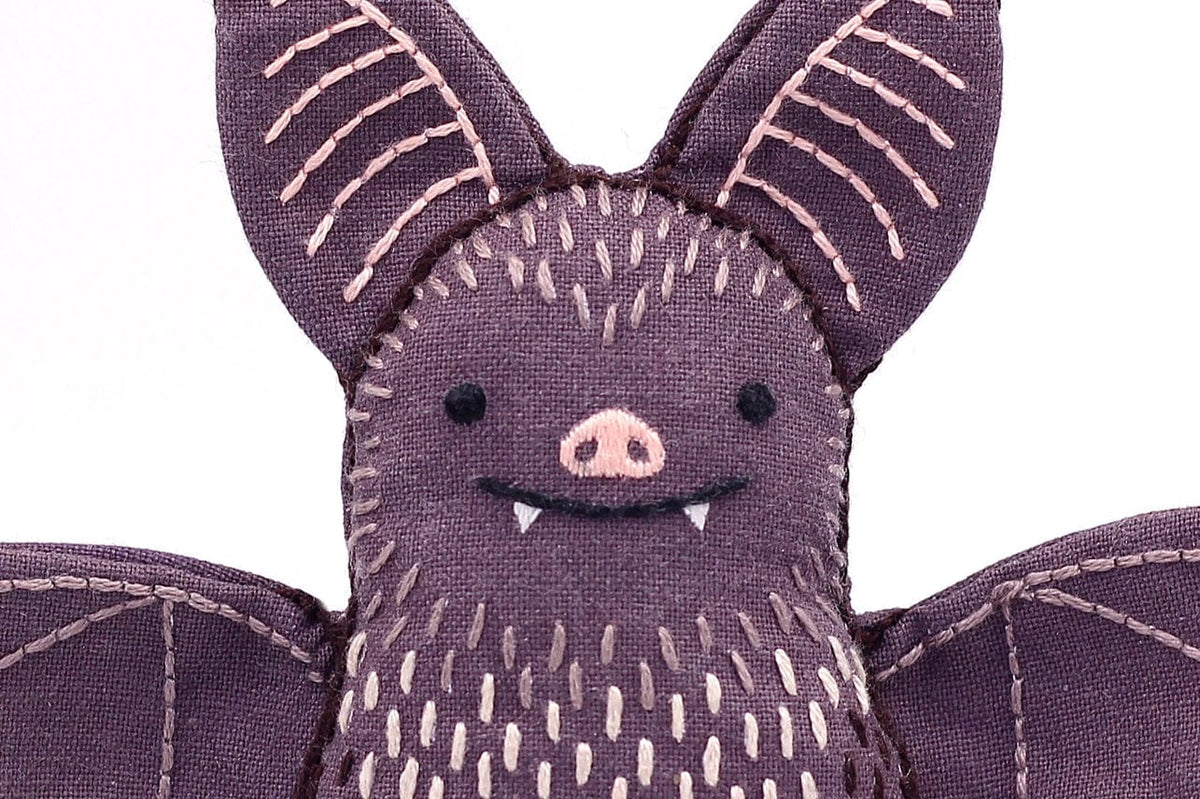 A close-up of the Kiriki Press Beginner Bat Embroidery Doll Kit shows a purple plush bat with stitched details, a smiling face with tiny fangs, and open wings—ideal for DIY embroidered doll projects.