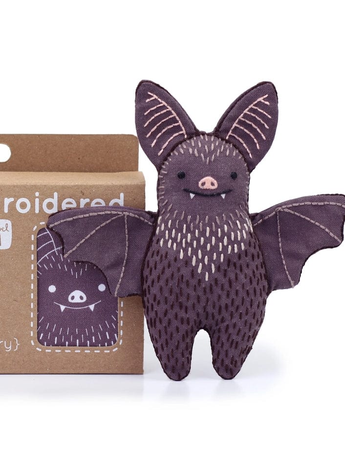 The Kiriki Press Beginner Bat Embroidery Doll Kit features a plush bat doll with embroidered details, large ears, and outstretched wings beside a brown box printed with the bat—ideal for fans of embroidery kits.