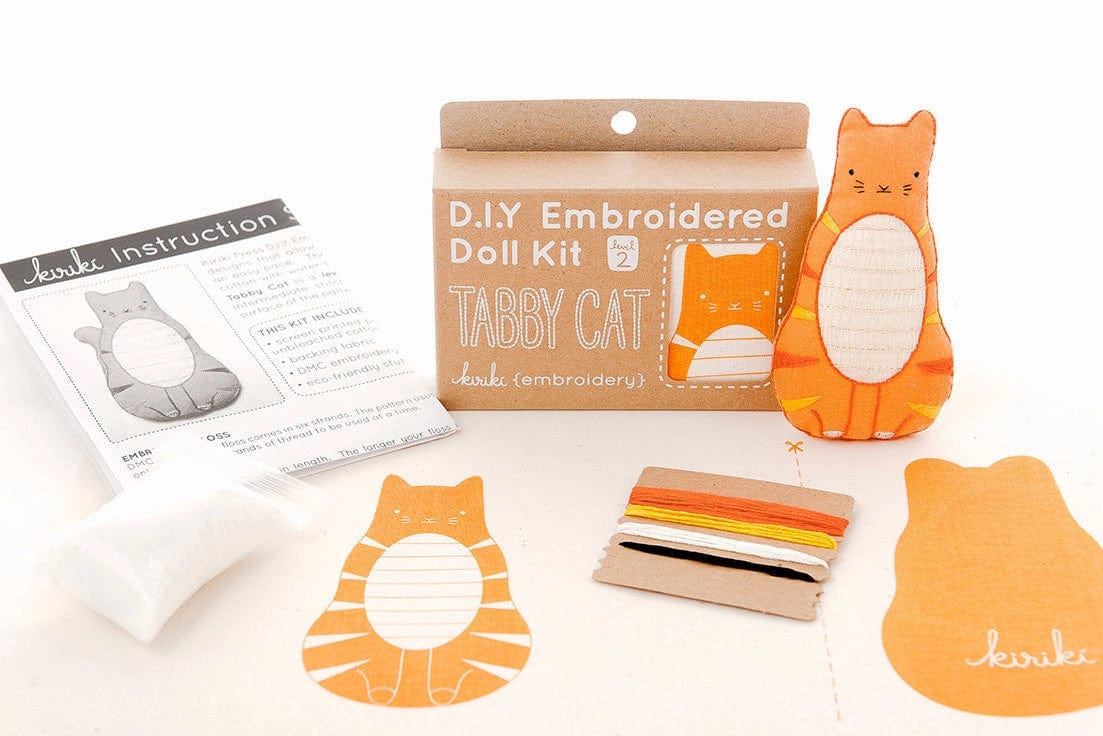 The Kiriki Press Beginner Tabby Cat Embroidery Doll Kit includes instructions, stuffing, colorful threads, a fabric piece with tabby cat outline, a cardboard bobbin, and a finished stuffed cat doll—perfect for first-time embroiderers.