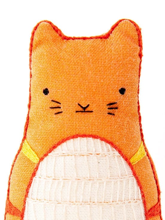 A close-up of Kiriki Press’s Beginner Tabby Cat Embroidery Doll Kit shows an orange plush with stitched black eyes, nose, whiskers, a white textured belly, and large ears—ideal for D.I.Y. embroidery fans.
