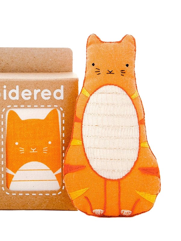 The Kiriki Press Beginner Tabby Cat Embroidery Doll Kit includes a plush orange tabby cat doll with a white belly and embroidered details, plus a matching illustrated brown box—ideal for creative beginners.