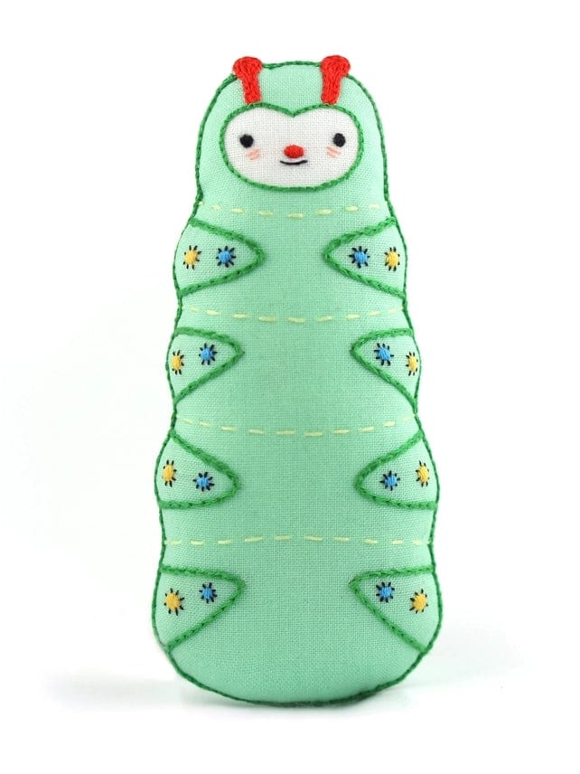 The Kiriki Press Beginner Caterpillar Embroidery Doll Kit features a soft, plush caterpillar with a cute face, red antennae, and green segments with blue and yellow flowers—ideal for creative DIY embroidery fun.