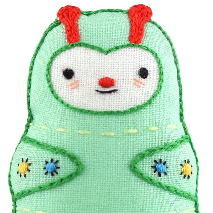 The Kiriki Press Beginner Caterpillar Embroidery Doll Kit features a handmade green fabric doll with red antennae, a white face, black eyes, pink cheeks, small smile, and blue/yellow embroidered arm details.