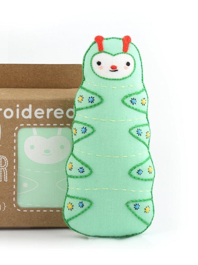 The Kiriki Press Beginner Caterpillar Embroidery Doll Kit features a soft green caterpillar plush with embroidered details, red antennae, and floral patterns—a perfect DIY starter. A matching illustration is shown on the brown box in the background.