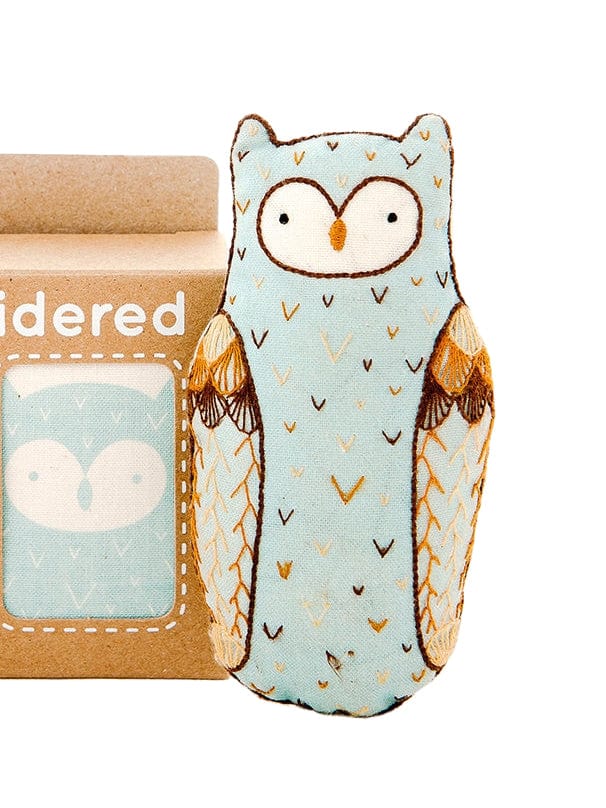 The Kiriki Press Beginner Horned Owl Embroidery Doll Kit includes a soft, light blue stitched owl doll displayed beside its brown box, which features a matching simple owl illustration on the packaging.