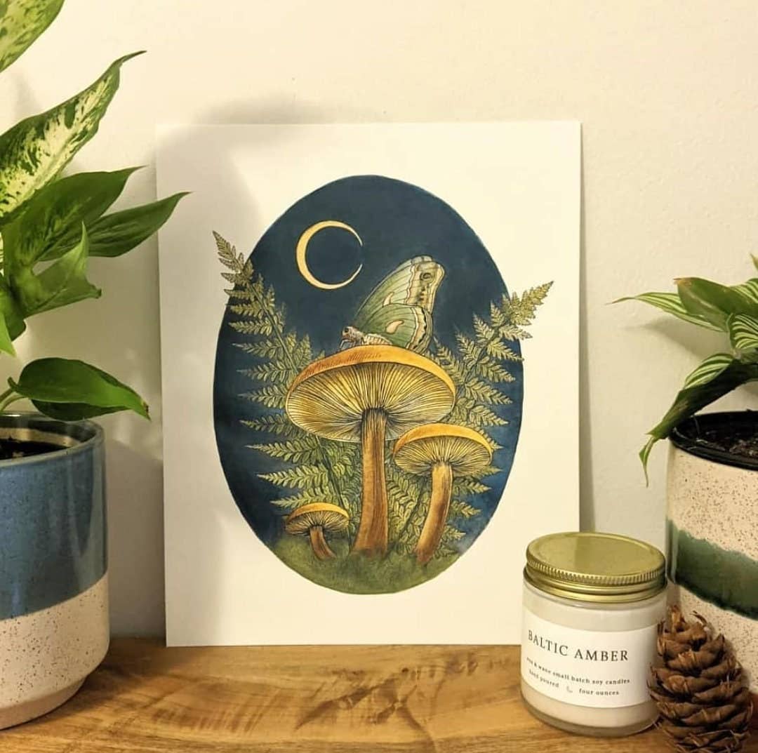 The Watercolor Mushroom Art With Moth and Fern Art Print by Kyla Rae Design (10 x 8) features woodland mushrooms, ferns, a butterfly moth, a crescent moon, potted plants, a pine cone, and a Baltic Amber candle on a cozy tabletop.