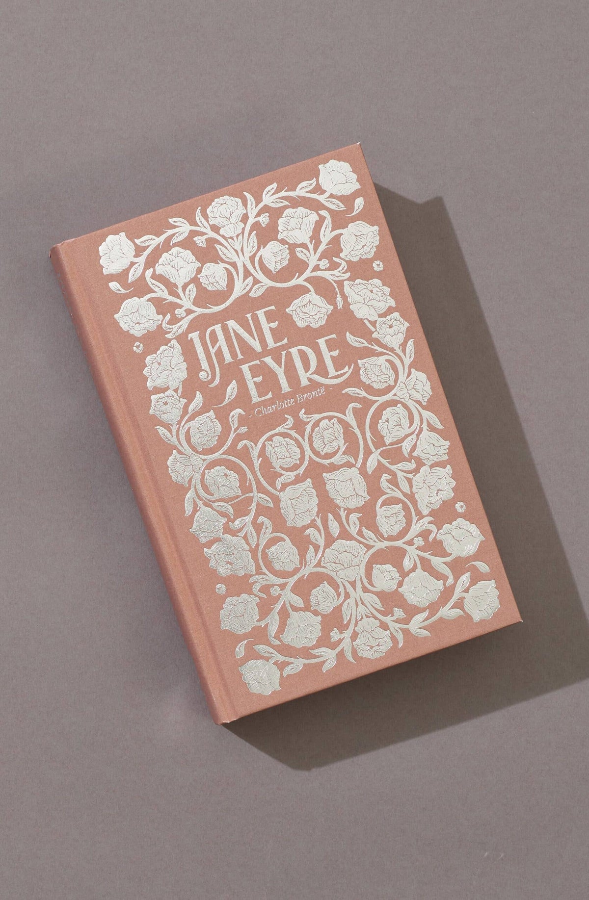 A pink hardcover edition of Jane Eyre by Charlotte Brontë from Wordsworth Classics, featuring an ornate silver floral design, is displayed on a neutral gray background—classic English fiction in sophisticated style.