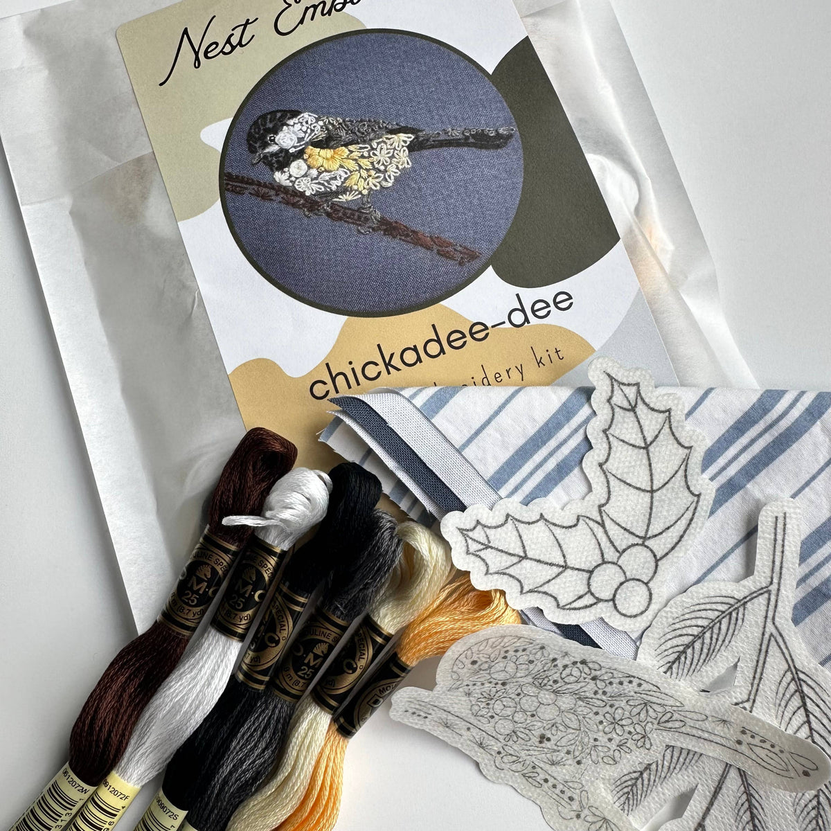The Nest Embirdery 4 Beginner Chickadee-dee Embroidery Kit includes colored threads, striped fabric, and easy patterns, featuring a stitched black-capped chickadee—ideal for bird lovers new to embroidery.