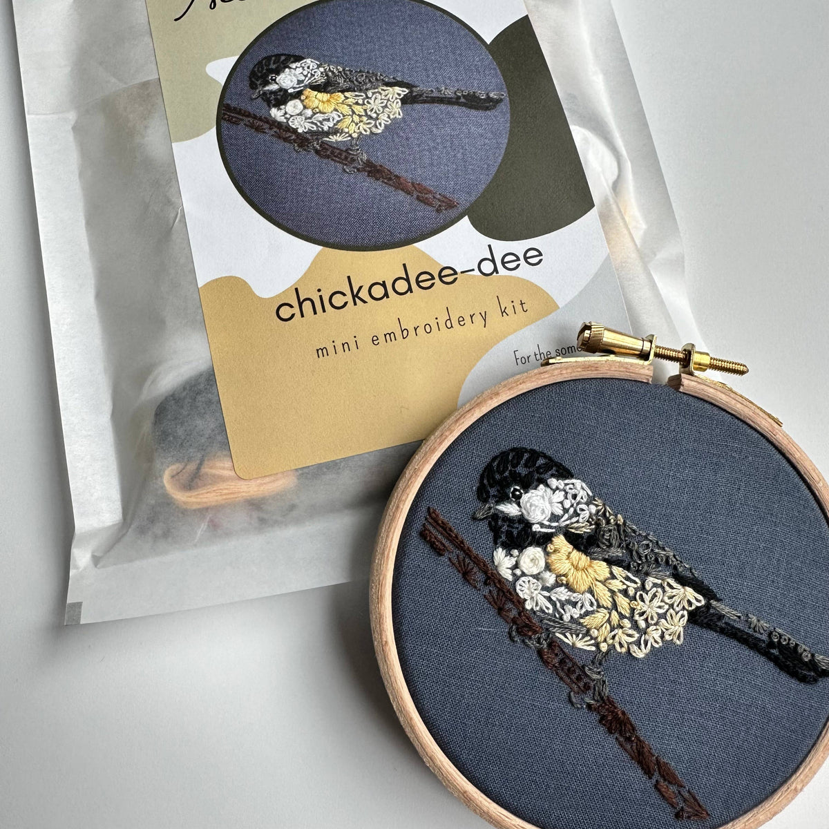 The Nest Embirdery 4 Beginner Chickadee-dee Embroidery Kit is shown next to a finished hoop, featuring a black-capped chickadee perched on a branch against dark fabric.
