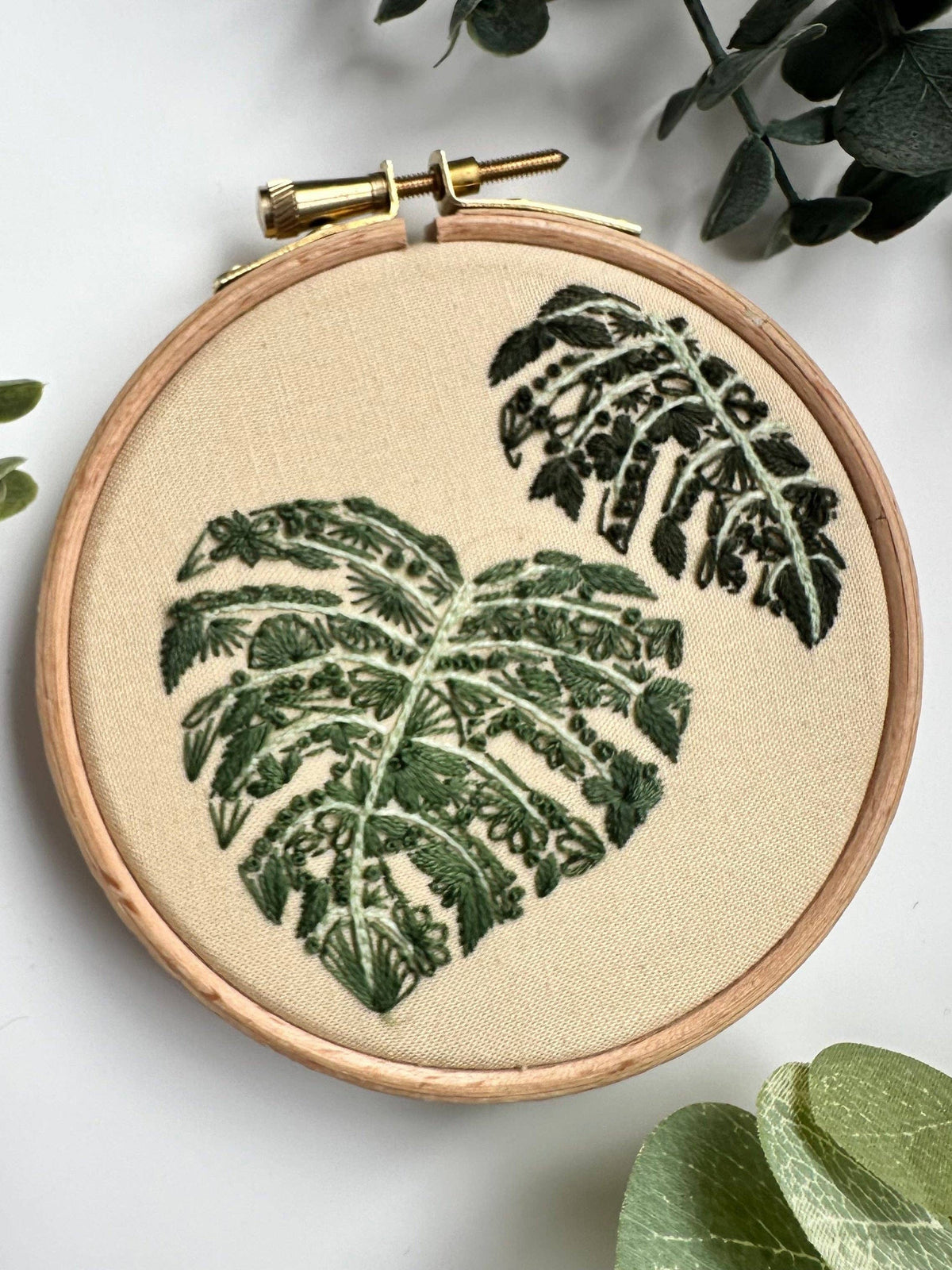 The Nest Embirdery 4 Beginner Mighty Monstera Embroidery Kit features two green monstera leaves stitched on fabric, surrounded by leafy accents—ideal for stick and stitch projects or as a mini embroidery kit.