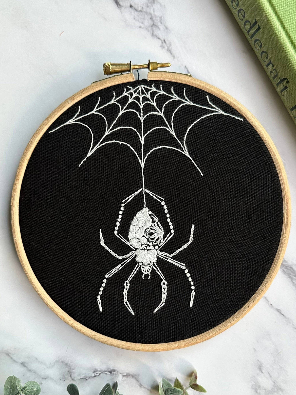 The Nest Embirdery 6 Beginner Orb Weaver Spider Embroidery Kit features a white orb weaver pattern on black fabric—ideal for embroidery beginners, with a detailed design and all you need to get started.