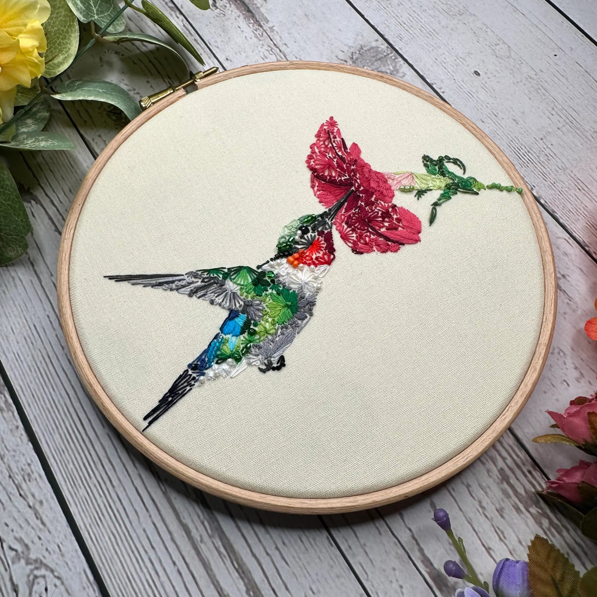 The Nest Embirdery 6 Spring Equinox Hummingbird Deluxe Embroidery Kit features a floral hummingbird sipping from a pink flower, beautifully displayed in a wooden hoop—ideal for Stick n Stitch designs or as a deluxe kit.