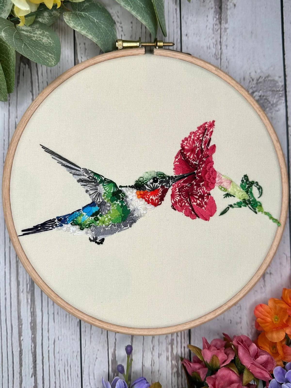 The 6 Spring Equinox Hummingbird Deluxe Embroidery Kit by Nest Embirdery features a vibrant floral hummingbird sipping nectar, displayed in a wooden hoop—an easy, beautiful project with Stick n Stitch designs.