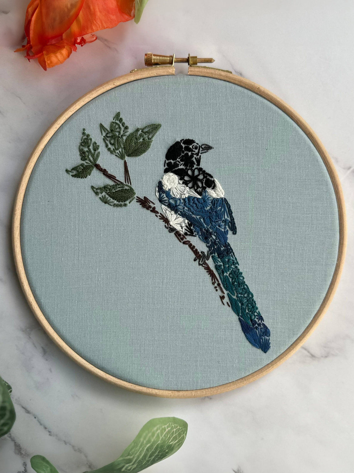 The Nest Embirdery 7 Beginner Mister Magpie Embroidery Kit features a detailed magpie perched on a leafy branch in black, white, and blue threads on light gray fabric, displayed on marble with orange and green plants nearby.
