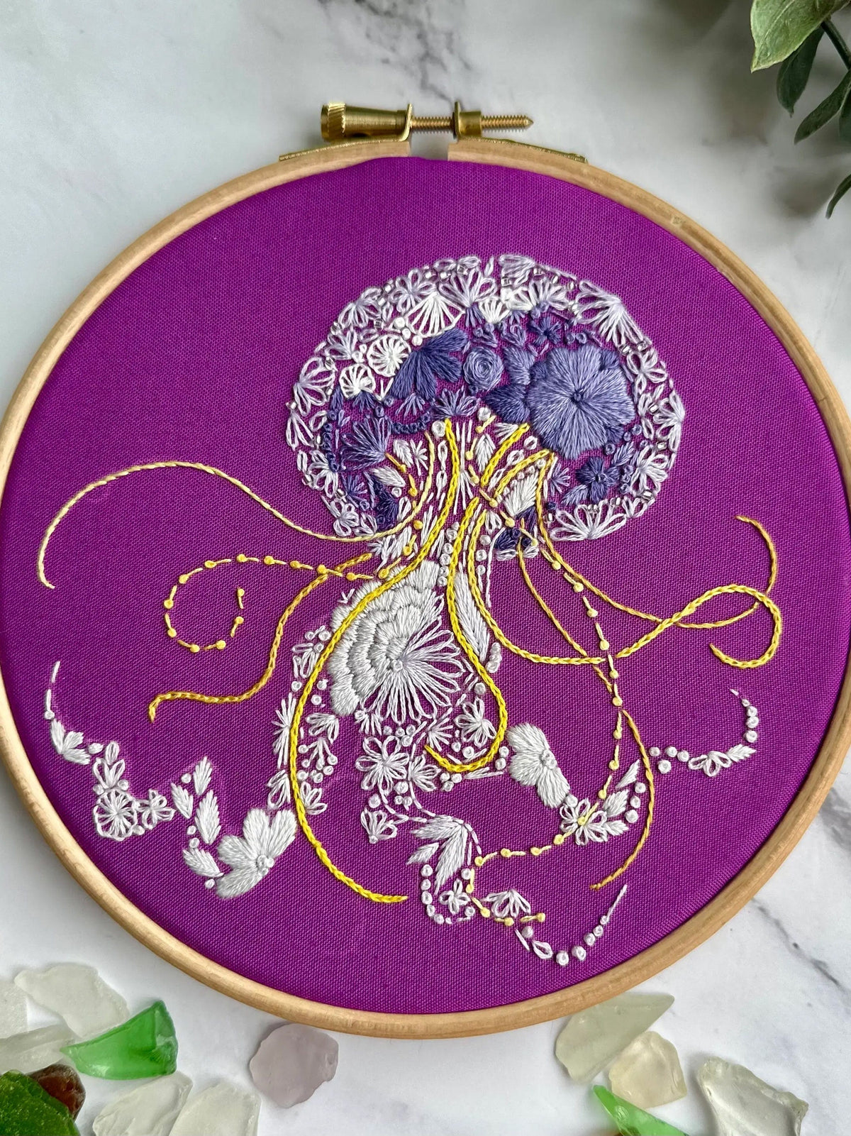 The 6 Beginner Jelly-Stitch Embroidery Kit by Nest Embirdery features detailed white, gray, and yellow threads on purple fabric in a wooden hoop, accented with green leaves and sea glass—perfect for beginner embroiderers.