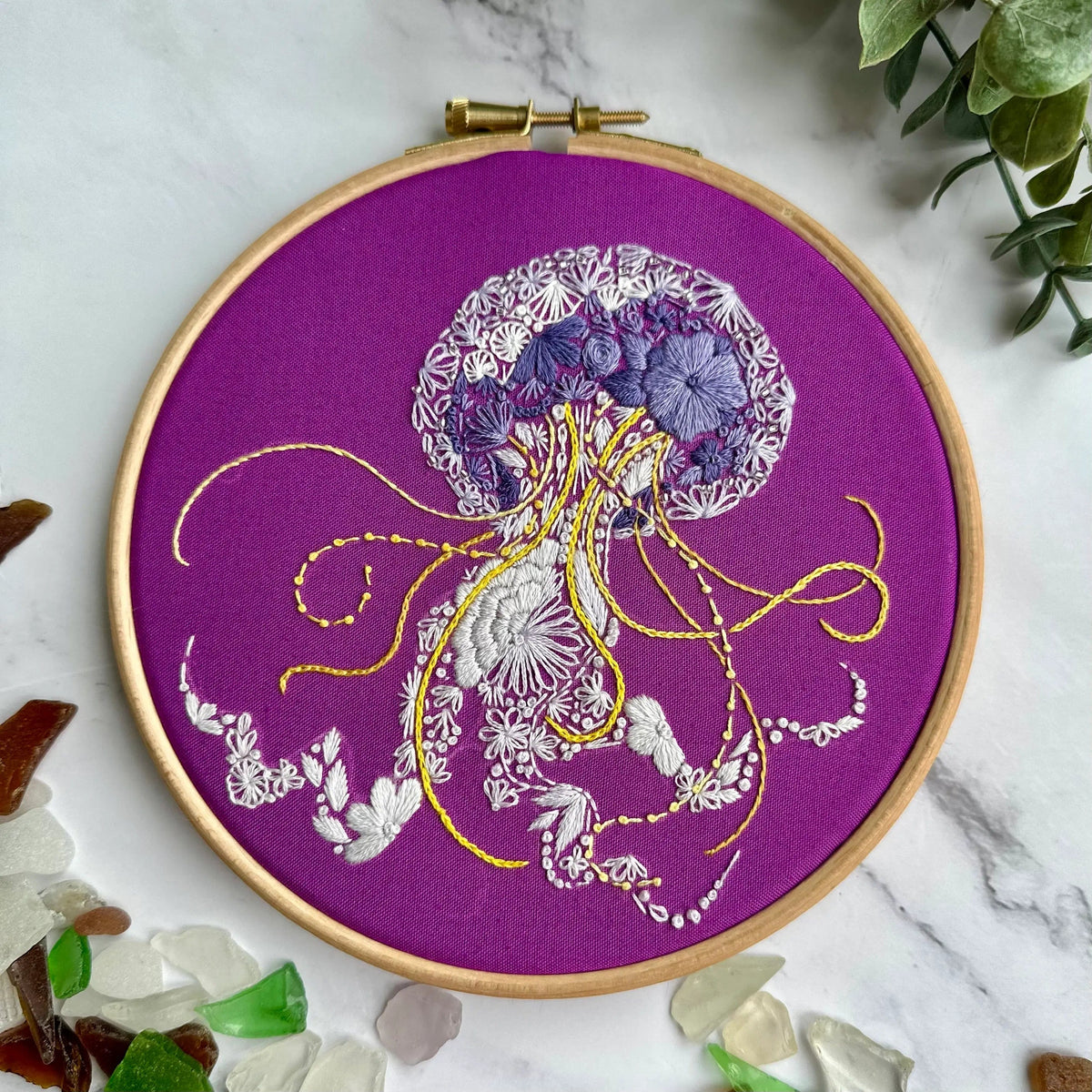 The Nest Embirdery 6 Beginner Jelly-Stitch Embroidery Kit features a purple hoop showcasing a jellyfish design stitched in white, yellow, and lavender with floral patterns on its body and tentacles, surrounded by glass pieces and green leaves.