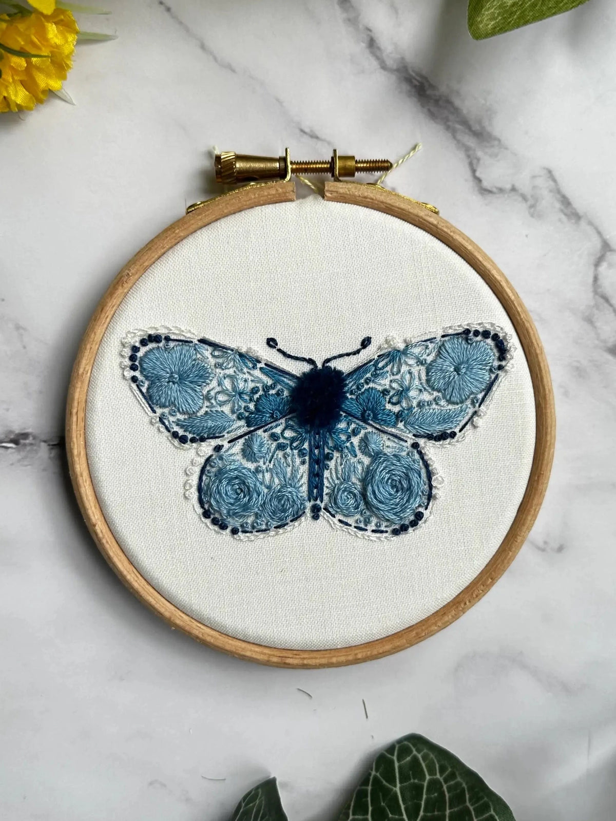 Nest Embirdery’s 4 Beginner Karner Blue Butterfly Embroidery Kit features a floral butterfly design on white fabric in a hoop, displayed on marble with green leaves and a yellow flower nearby.