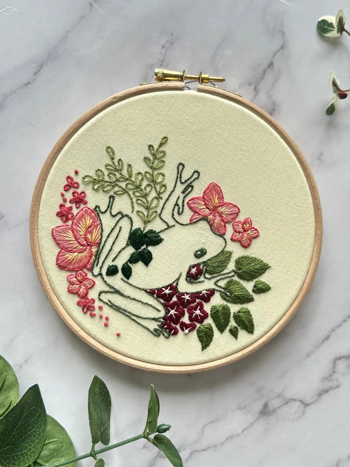 The Nest Embirdery 6 Beginner Froggy Flowers Embroidery Kit features a stitched white tree frog among green leaves and pink flowers on cream fabric—ideal for advanced beginners ready for a fun, floral project.