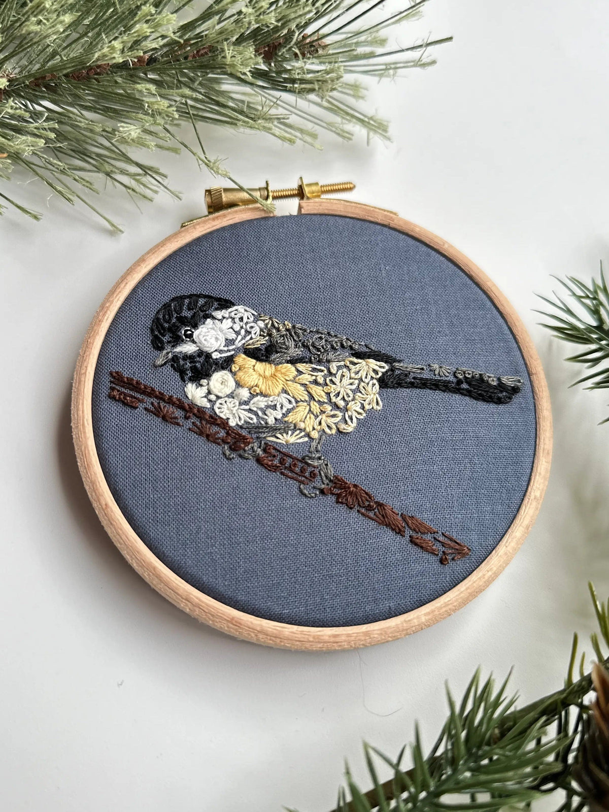 The Nest Embirdery 4 Beginner Chickadee-dee Embroidery Kit features a hand-stitched black-capped chickadee on blue-gray fabric with pine needles—perfect for bird lovers looking to try embroidery.