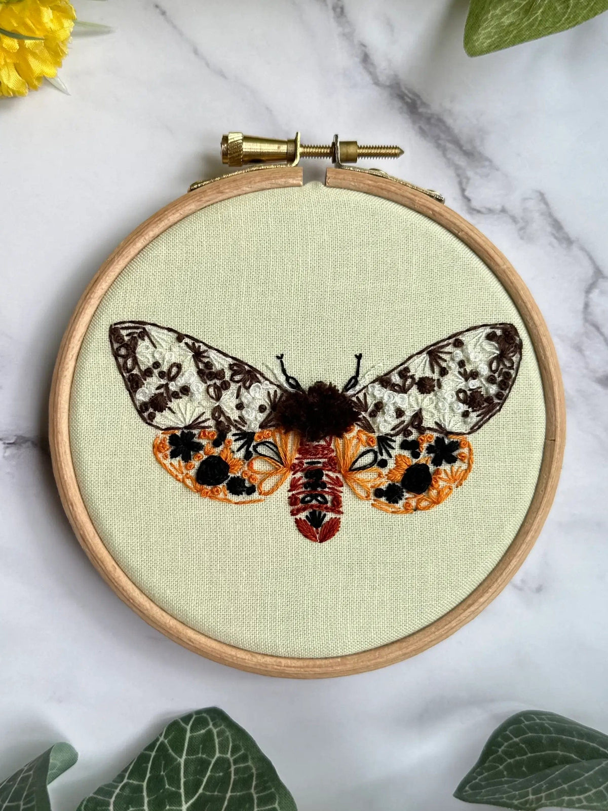 The 4 Beginner Garden Tiger Moth Embroidery Kit by Nest Embirdery features a wooden hoop holding fabric embroidered with a colorful brown, black, orange, and white moth amidst green leaves and a yellow flower on marble.