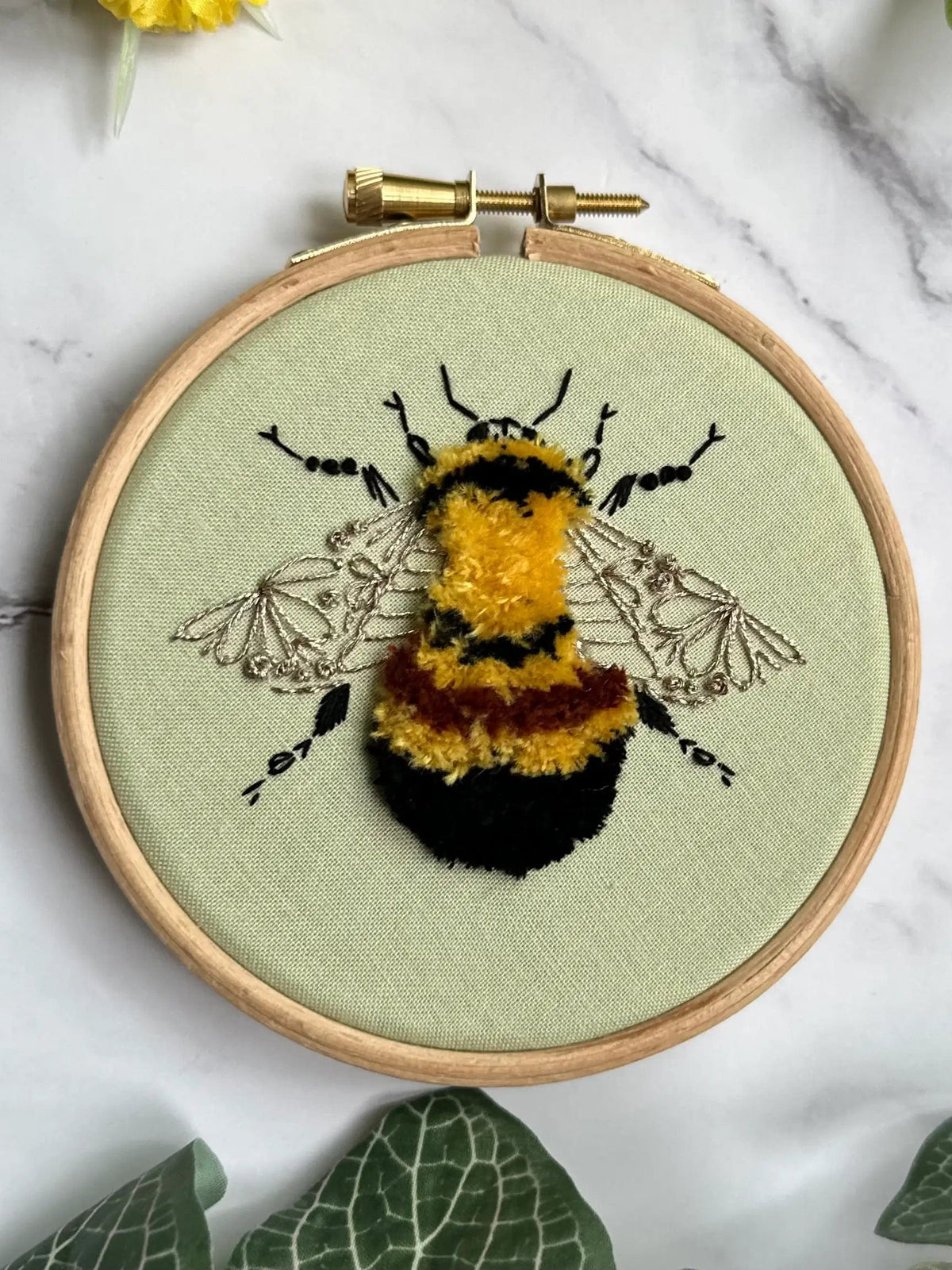 The Nest Embirdery 4 Beginner Rusty Patched Bumblebee Embroidery Kit features a textured bee in yellow, orange, and black threads with black-outlined wings on pale green fabric—perfect for DIY embroidery projects.