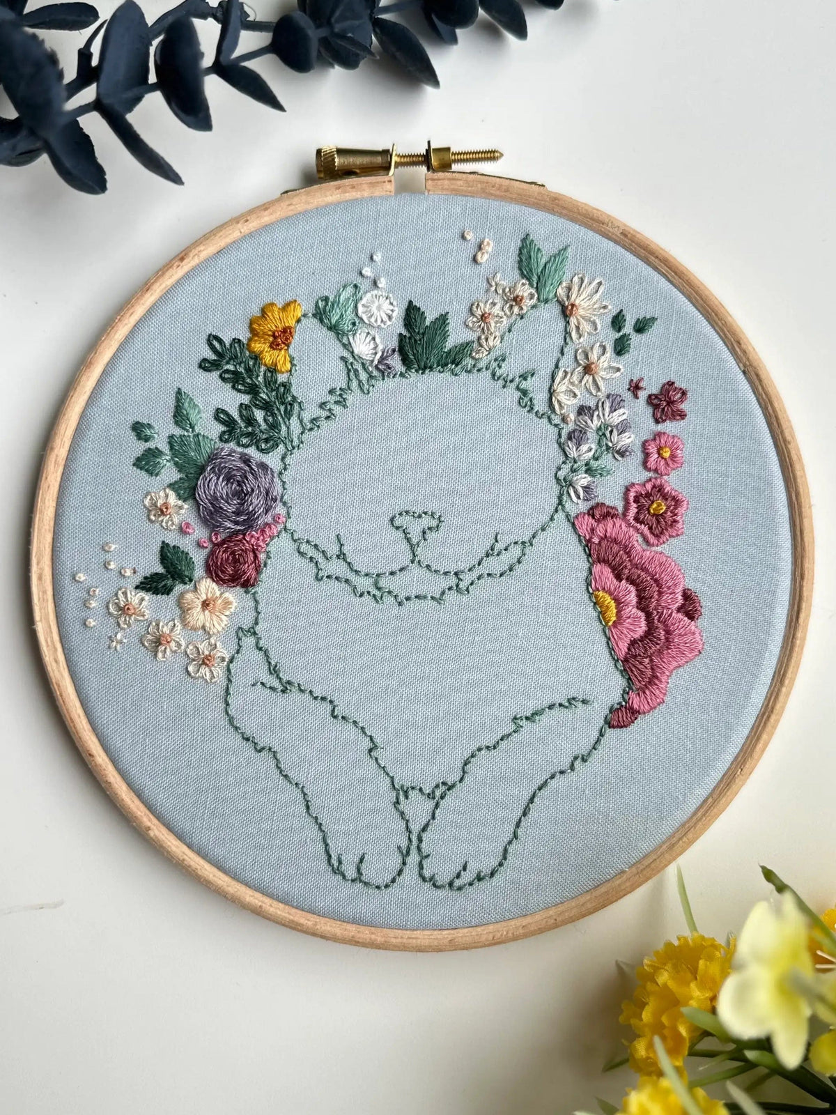 The Nest Embirdery 6 Beginner Kitty Bouquet Kit is ideal for advanced beginners, featuring a simple green kitty outline surrounded by colorful embroidered flowers in yellow, pink, purple, and white on light blue fabric.