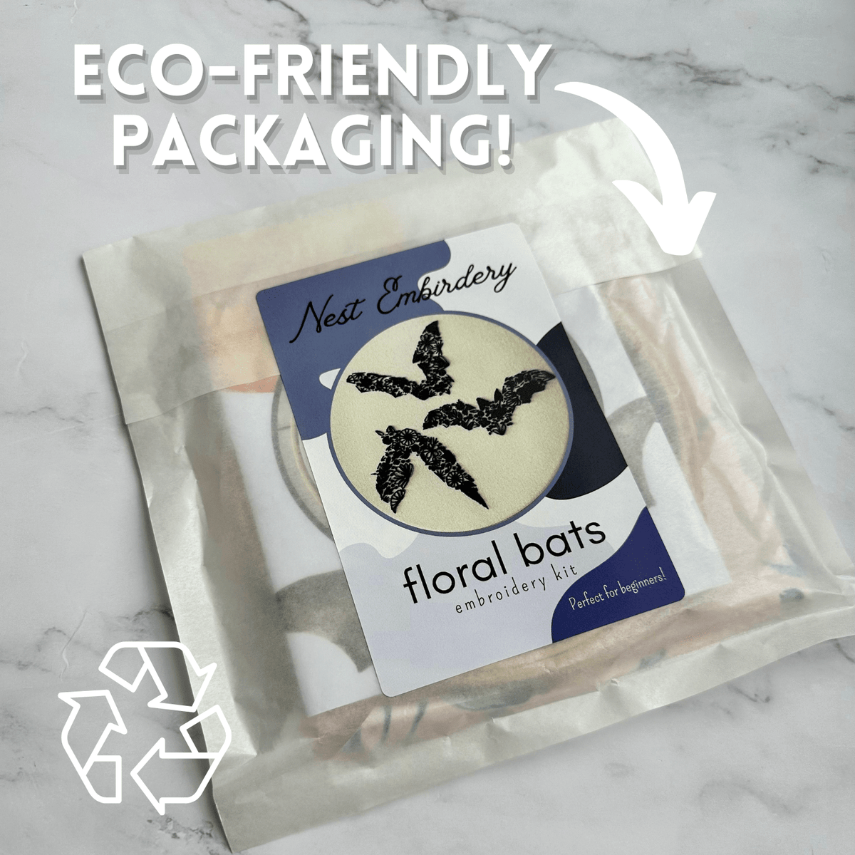 Nest Embirderys *Late May Pre-Order* 6 Beginner Floral Bat Embroidery Kit is in a translucent, eco-friendly package on marble. Text reads ECO-FRIENDLY PACKAGING! with a recycling symbol and white arrow pointing at the package.