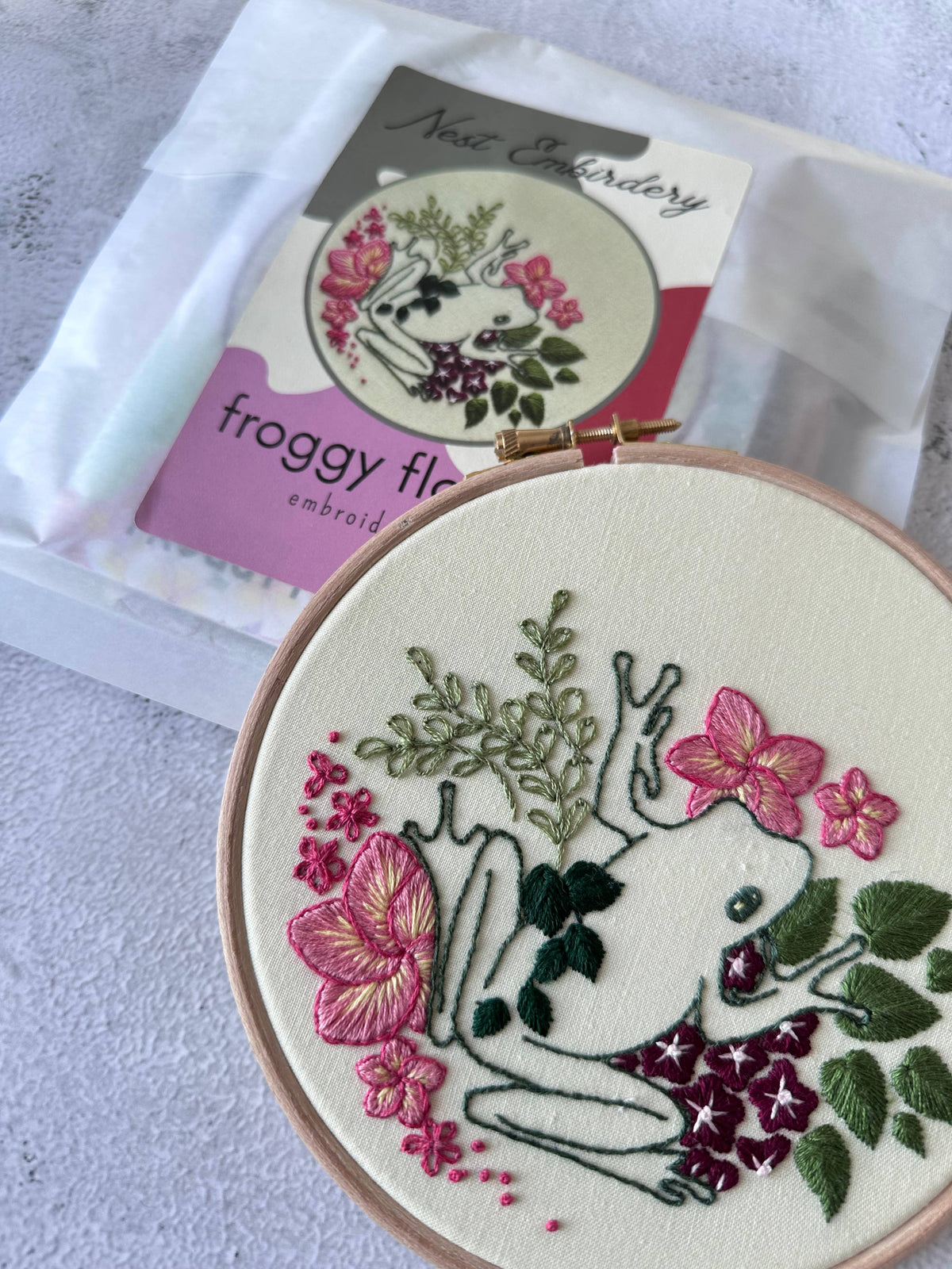 A 6 hoop by Nest Embirdery shows a white frog among pink flowers and green leaves. The same froggy design appears on the 6 Beginner Froggy Flowers Embroidery Kit packet—perfect for advanced beginner embroiderers.